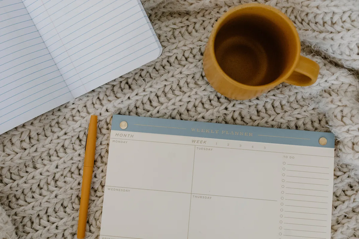 A weekly planner laying on a bed with pen and coffee mug