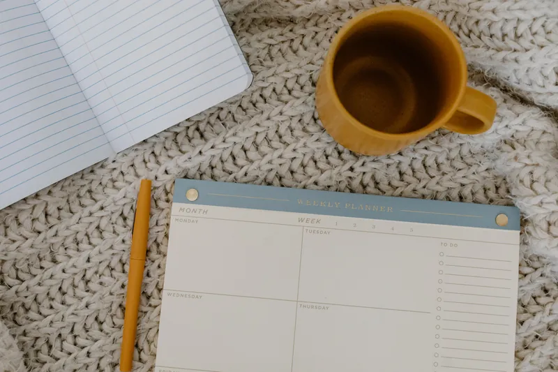 A weekly planner laying on a bed with pen and coffee mug